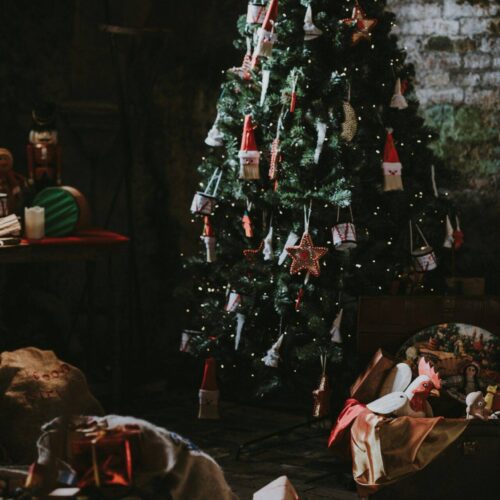 annie-spratt-1Qg9PXse00s-unsplash A decorated Christmas tree stands indoors with gifts, toys, and wrapped presents on the floor around it in a dimly lit room.