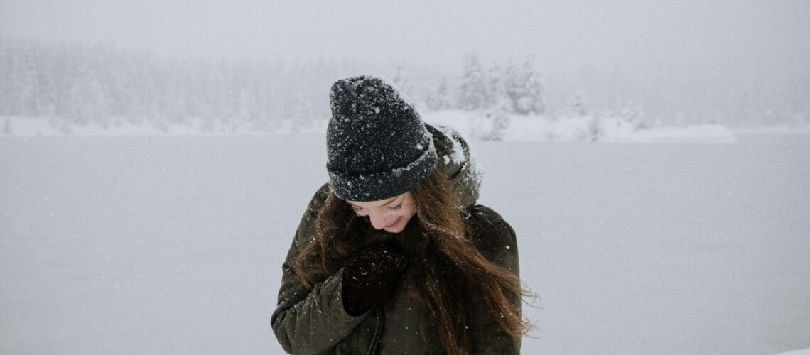 A person in a winter coat and beanie stands in a snowy landscape, looking down and smiling.