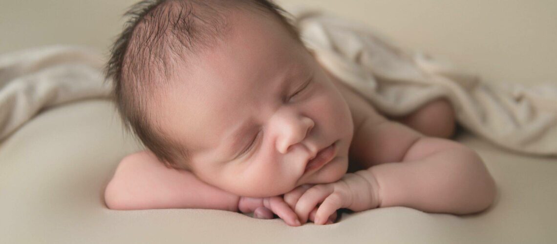 Newborn baby sleeping peacefully on a soft beige surface with a light blanket partially covering their back.