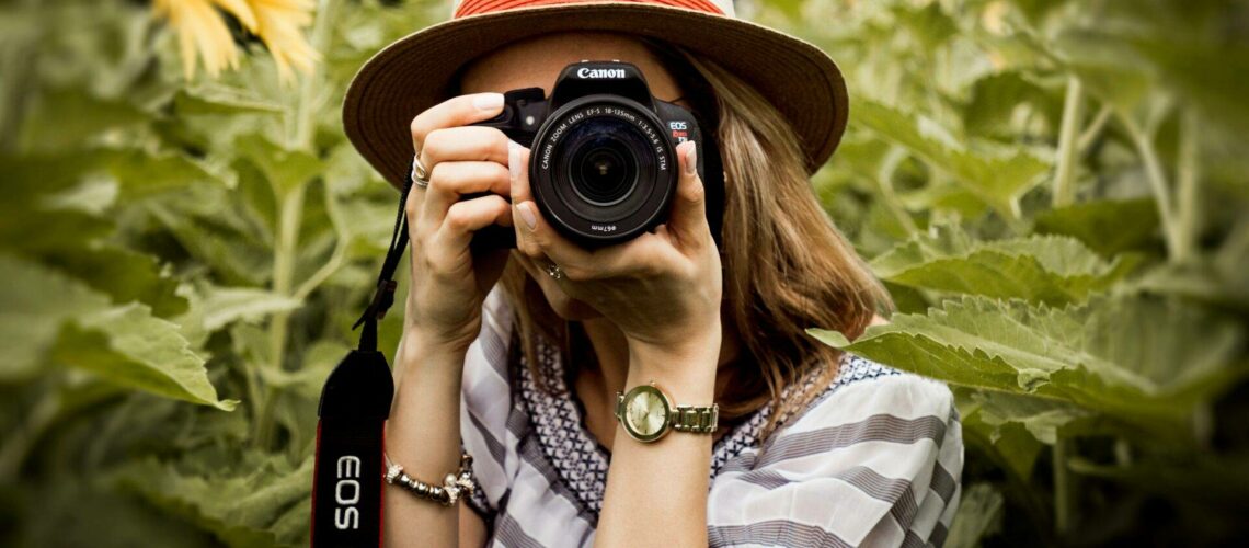 Person wearing a hat takes a photo in a sunflower field with a Canon camera.