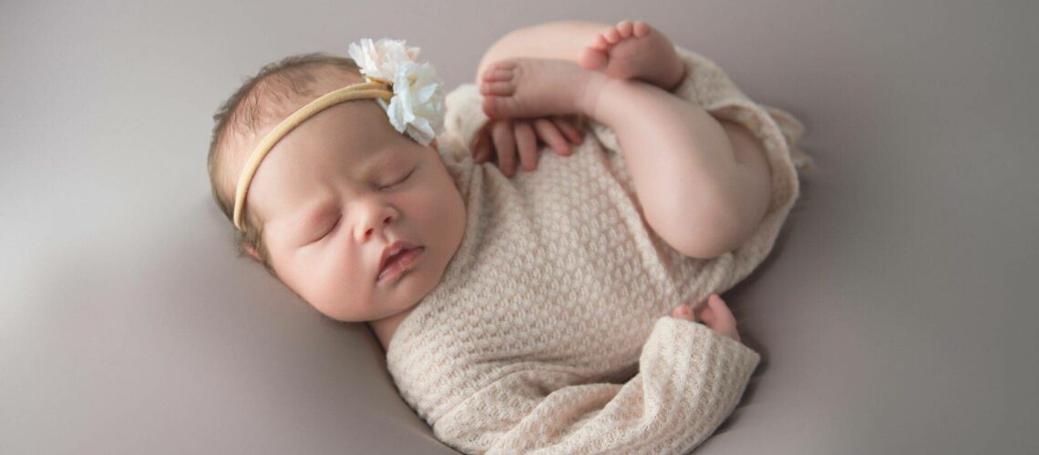 Sleeping newborn in a knitted outfit with a floral headband, lying on a soft, neutral-toned surface.