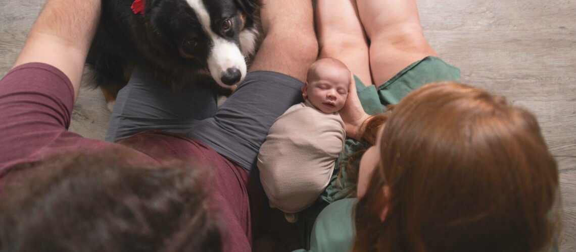 A baby wrapped in a blanket sleeps between two adults sitting on the floor. A dog with a red bow sits nearby, looking up.