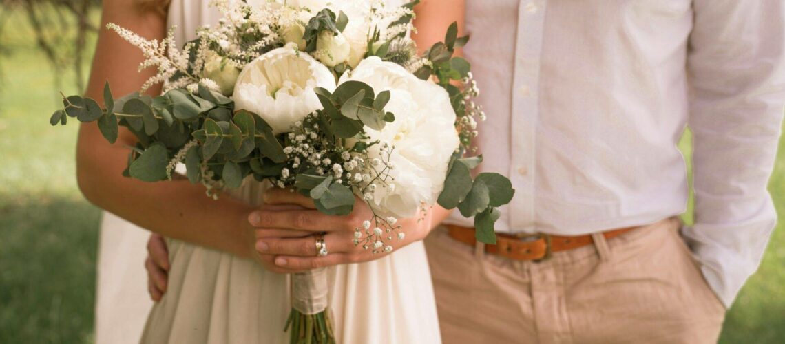A couple stands together, the woman holding a bouquet of white flowers with greenery. They are dressed in light, casual attire, and are outside with grass in the background.