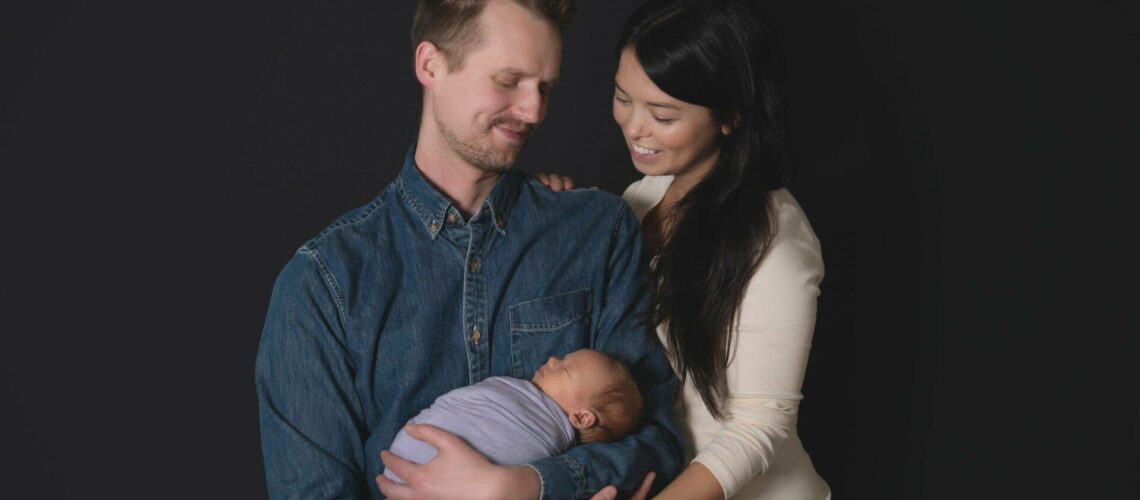 A seated man holds a sleeping baby in his arms, dressed in a blue onesie. A standing woman in a white top is beside him, leaning in with a smile. The background is black.