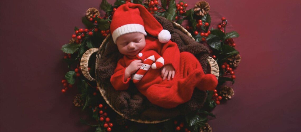 A newborn baby in a red outfit and Santa hat sleeps in a basket surrounded by holly leaves, red berries, and pine cones, set against a red background.