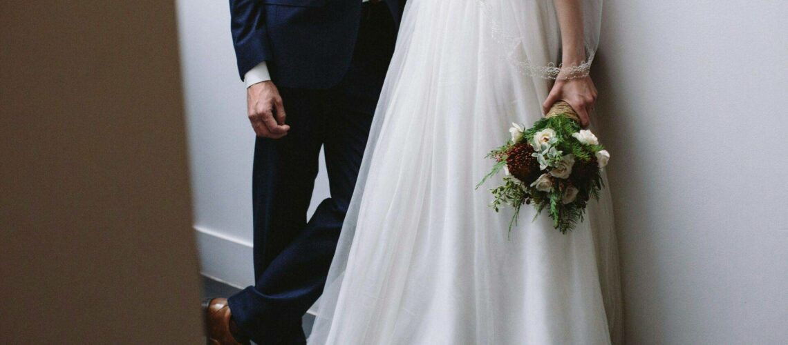 A bride and groom stand closely, leaning against a wall. The bride holds a bouquet of flowers. The groom is in a dark suit and brown shoes.