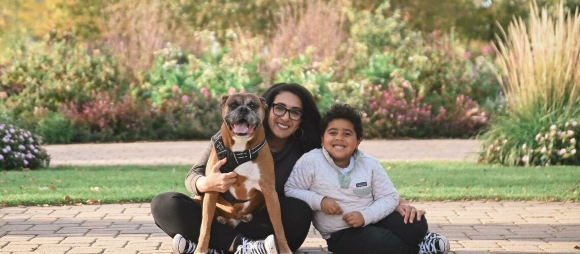 A person, child, and dog sit on a brick path in a park. The child and dog wear matching clothes. Greenery and trees are in the background.