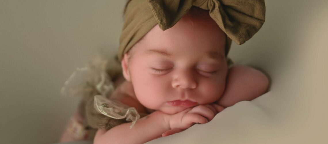 A sleeping baby with a large brown bow headband rests their head on folded arms.