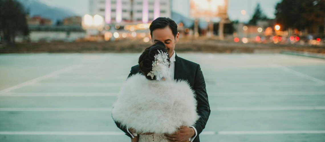 A couple embraces outdoors in formal attire, with the woman wearing a white dress and fur shawl. The background features a blurred building with lights.