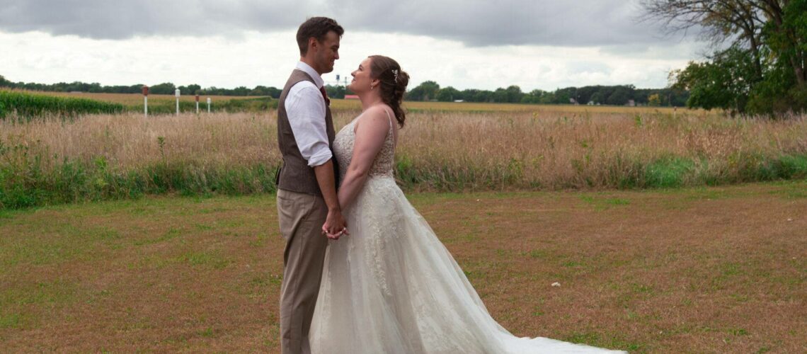 A bride and groom stand holding hands and facing each other in an open field, under a cloudy sky.