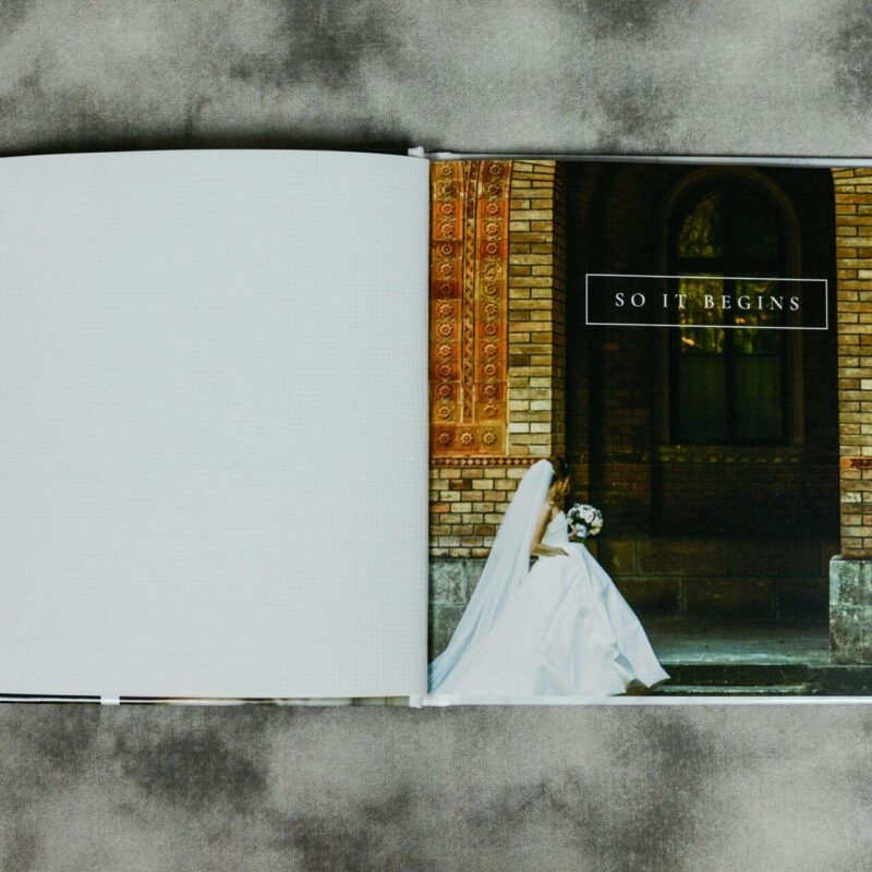 An open photo album shows a bride and groom on the right page with the caption "So It Begins" against a brick archway, the left page is blank.