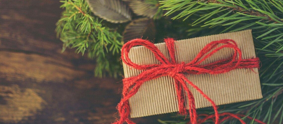 A small, wrapped gift with a red string bow rests on pine branches with a wooden background.