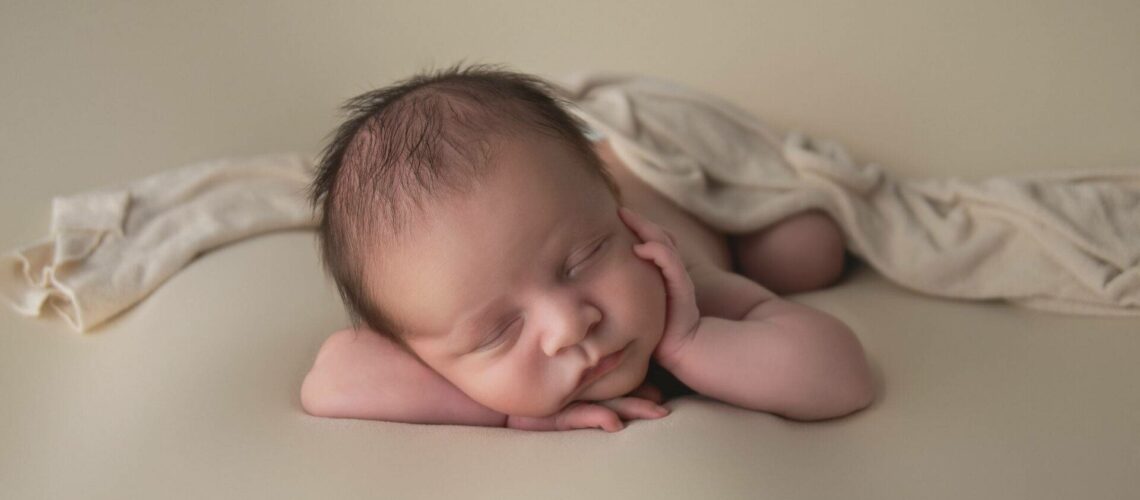 A sleeping baby rests with its head on its hands and a light cloth draped over its back, set against a soft beige background.