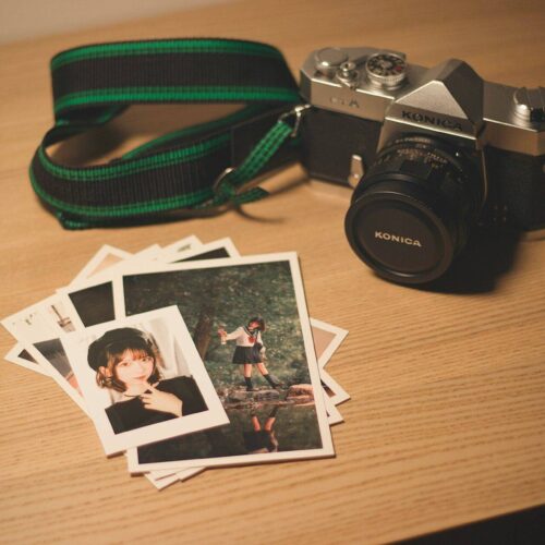 A Konica film camera with a green strap sits on a wooden surface next to several printed photographs, including portraits and an outdoor scene.